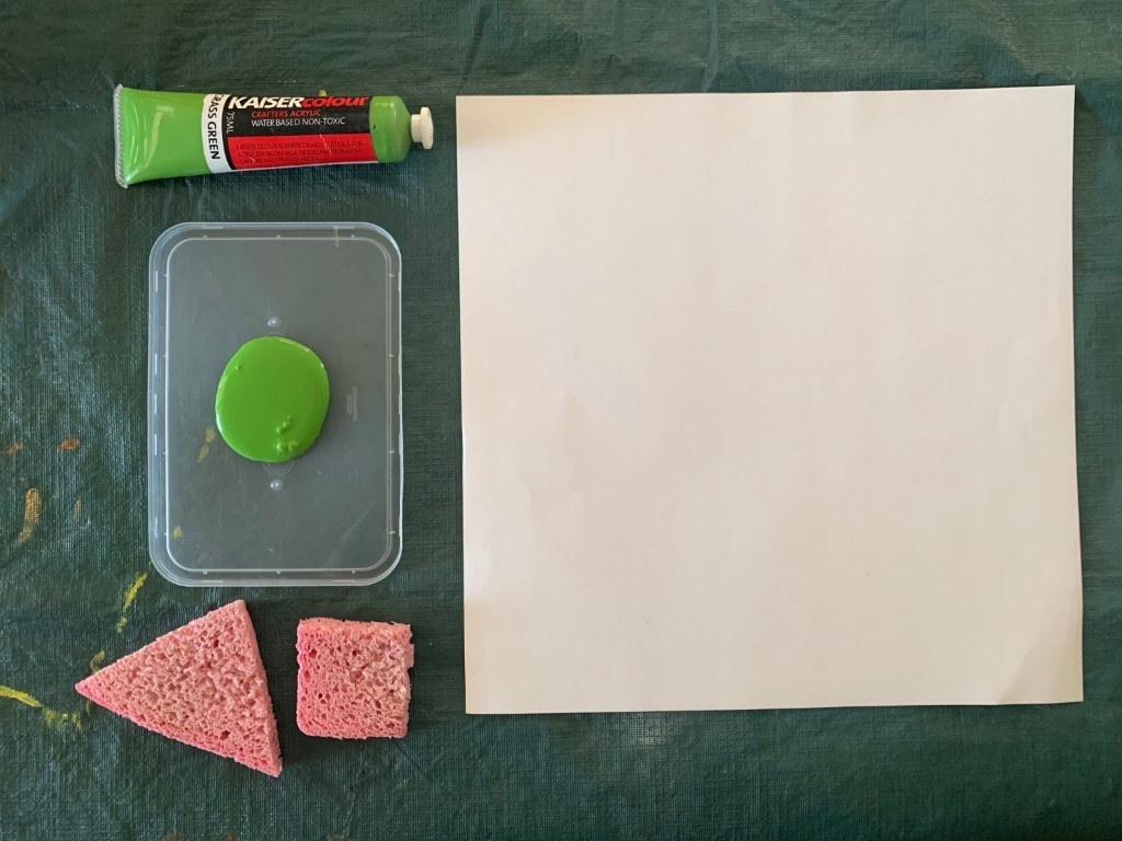 Image of how to set up for sponge art activity. It shows a tube of green paint, a plasctic lid with green paint squeezed onto it,  2 sponges and a blank piece of paper.