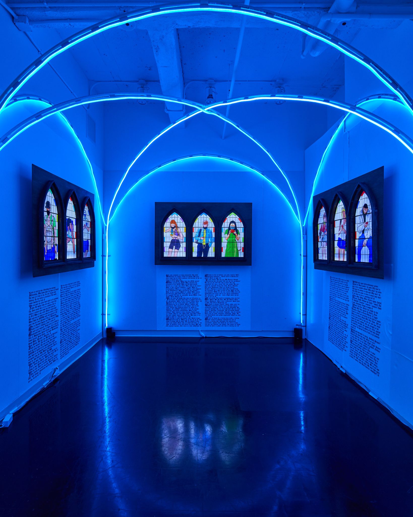 A small dark room with neon blue arches on the ceiling. On each wall are three small panels of stained glass with images of people in masks looking at their phones. Below each panel is a large block of text.