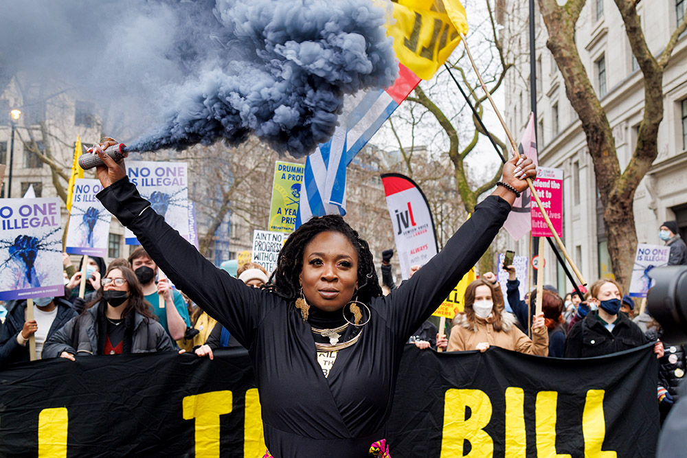 A black woman stands defiant holding a smoke flare at the head of a long rally though central London. A big banner says Kill The Bill.