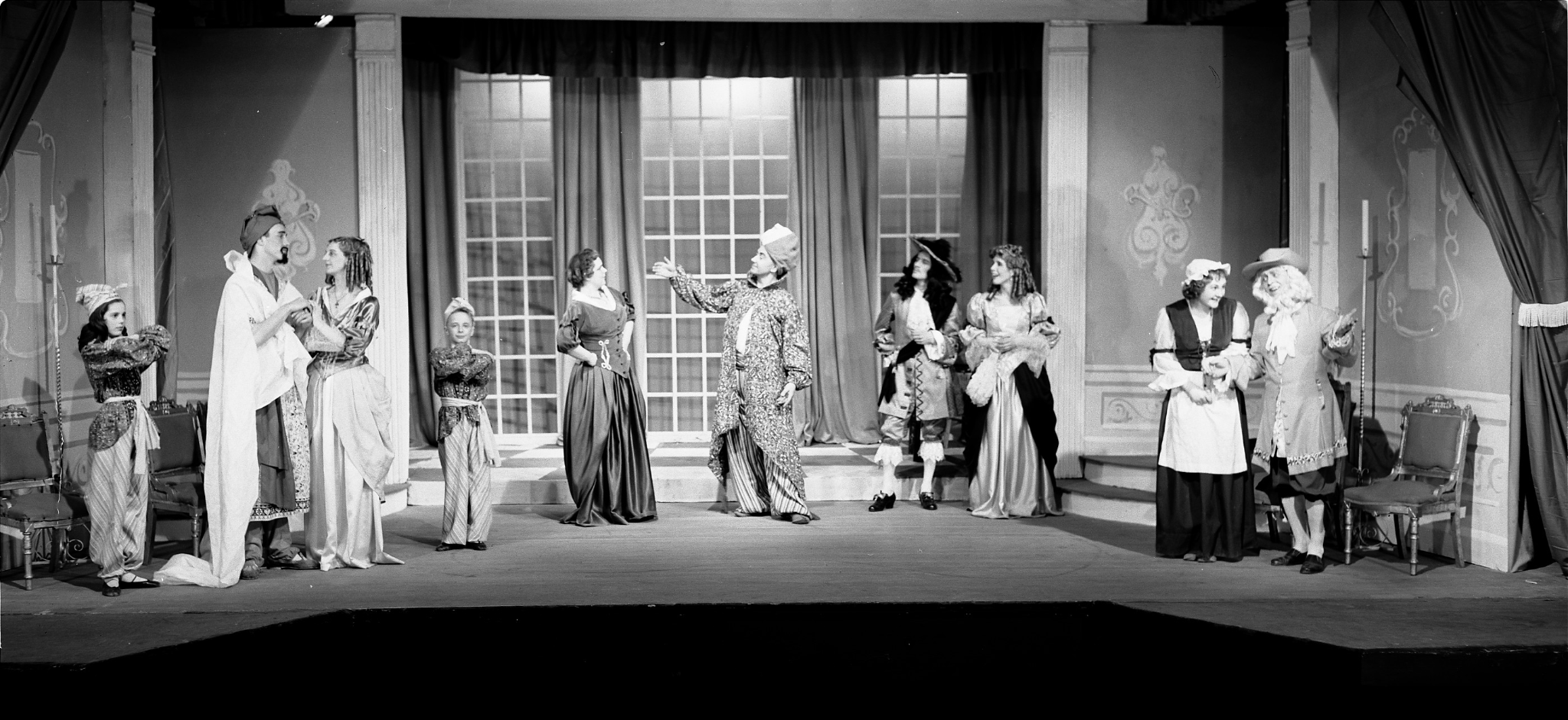 A cast of ten Caucasian actors stand on a stage designed to look like a grand ballroom. They wear seventeenth century clothing and stand in small conversation groups.