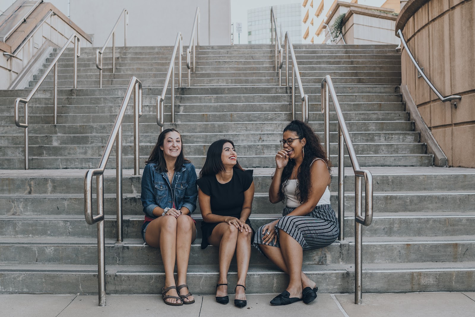 Tres chicas de entre los 25 y 30 años, sentadas en el segundo escalón más bajo de unas escaleras. Visten juvenil y para un clima cálido. Tiene una conversación y ríen