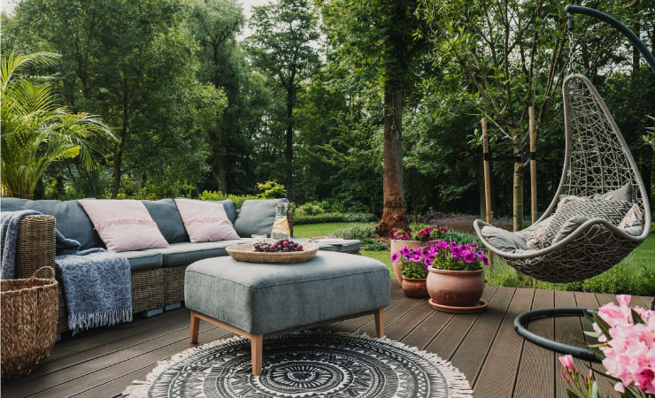 A chic outdoor patio with several design elements. There is a wicker sofa with complimentary cushions and throw pillows. There’s a round outdoor rug, with fringe all around it, an ottoman, and a hanging chair. 
