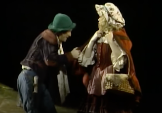 A man in a green hat is crouched down to speak to an actress playing a young girl with blonde, curly hair and a doll-like outfit.
