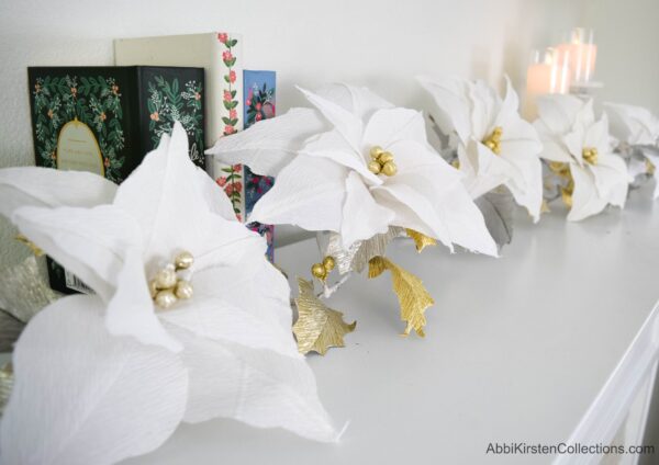 Giant white crepe paper poinsettia garland on a white mantel, with garland books and white candles. You can make this paper poinsettia for your Holiday celebration. 