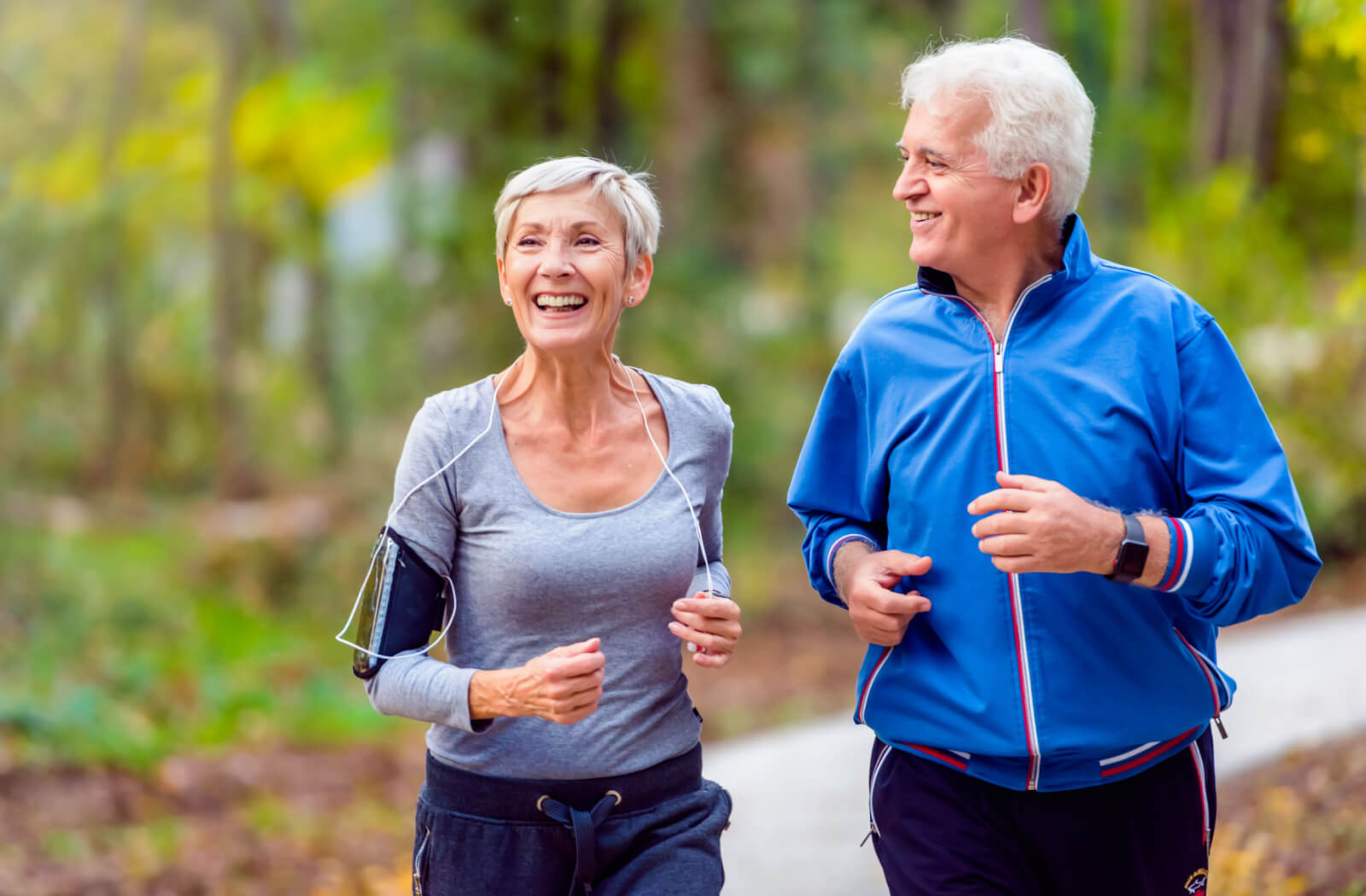 A senior woman and a senior man smiling while jogging in a park.