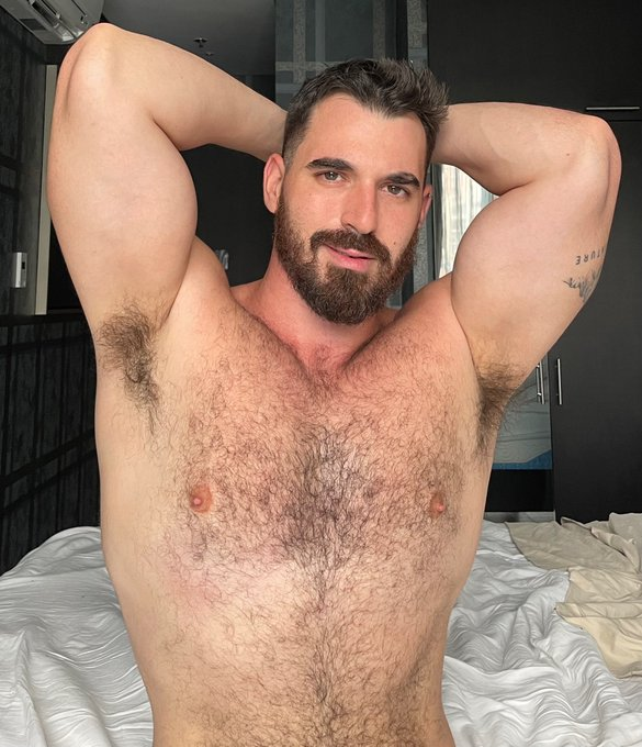 Thick Macho shirtless and smiling for the camera with his arms behind his head and sitting on white bed sheets in his bedroom