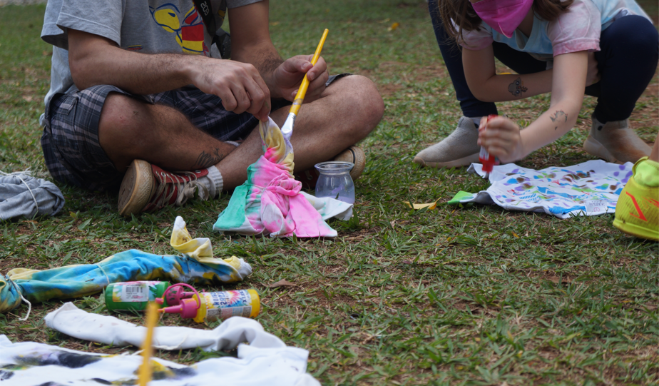 A imagem mostra pessoas sentadas na grama. Ao redor espalhados material de arte e tecidos.