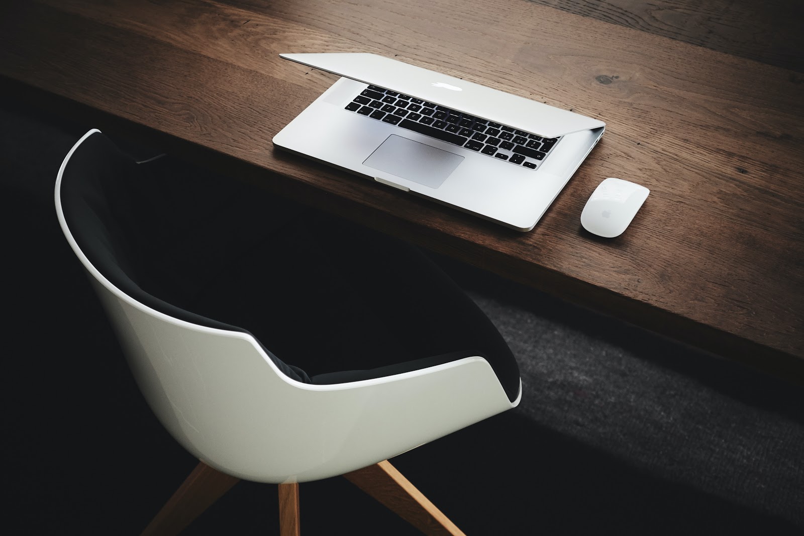 Imagem de uma mesa de madeira escura, com um notebook quase fechado, com um mouse sem fio ao lado. Junto à mesa, encontra-se uma cadeira com encosto e acento pretos, revestida por um material branco, com pés de madeira. O chão do local é preto.