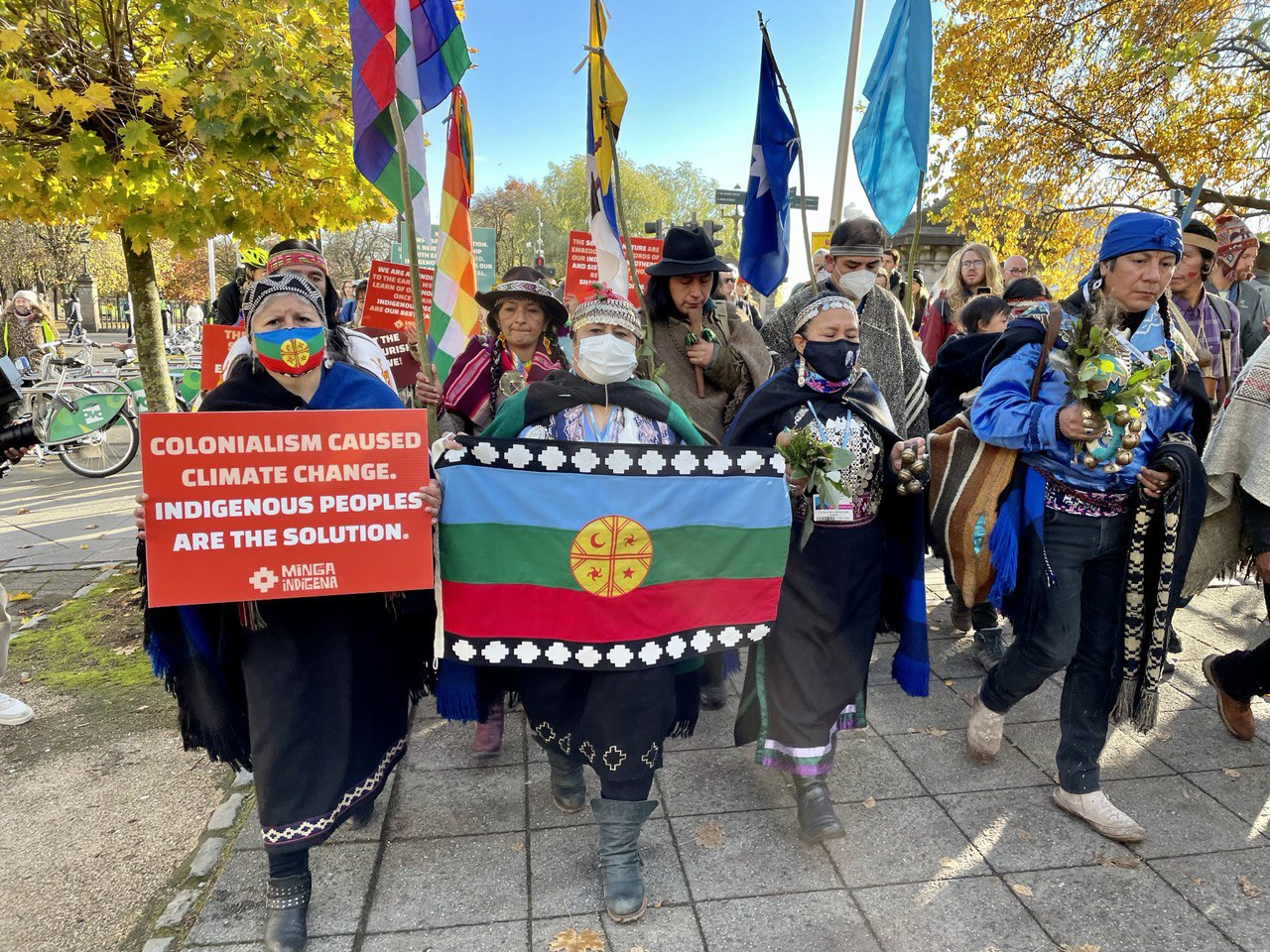 Indigenous people in beautiful dress hold banners and march towards camera.