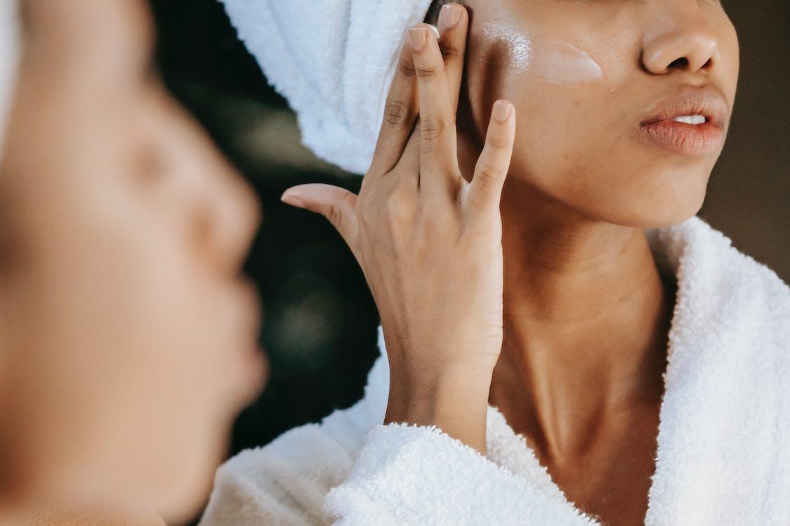 Free Crop unrecognizable young ethnic female in terry robe applying moisturizing cream on cheek while reflecting in mirror Stock Photo