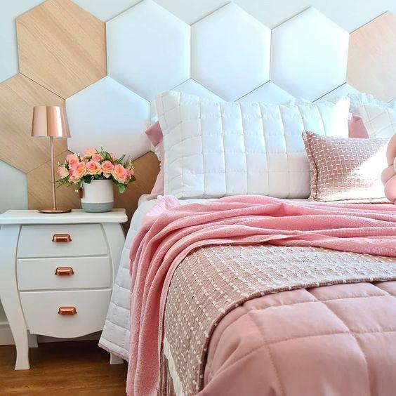 Quarto com cama de casal com cabeceira estofada em formatos hexagonal, amadeirado, branco e rosa, criado mudo branco com abajur e piso de madeira