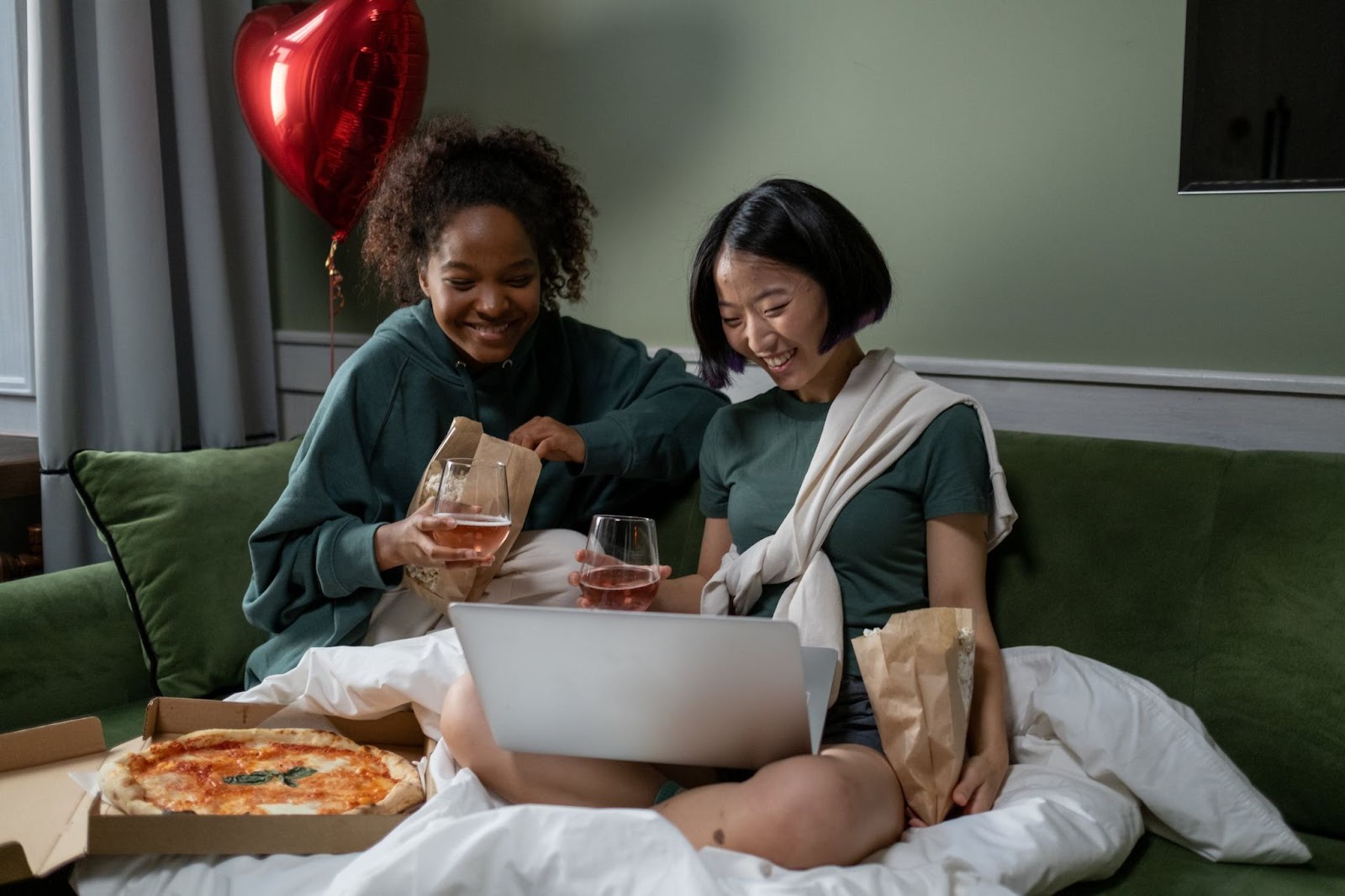 Two women drinking and eating on bed while watching a movie on a gray laptop 