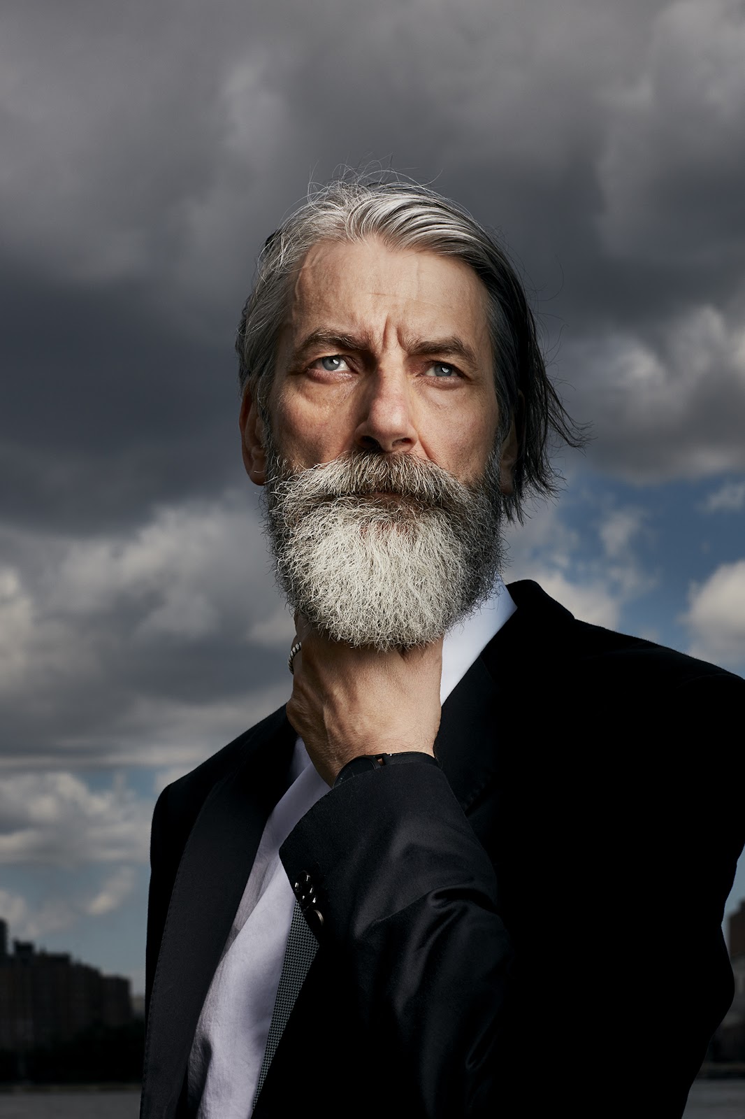 This moody image was created over the Covid-19 lockdown by Matt Carr as part of a personal project. The subject is a man in a suit looking very contemplative. He is fscing the camera, staring off into the distance. His silver hair and beard are wind-swept. In the background, gray, rain-heavy clouds cover the sky.