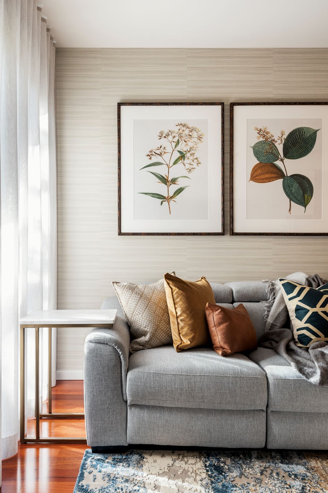 Living room with a light gray sofa, decorative throw pillows in warm tones, and framed botanical prints on the wall.