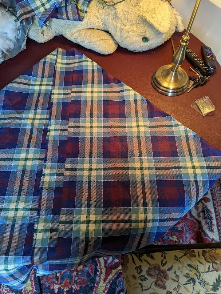 A piece of tartan fabric in blue, purple, green, cream, and silver is shown folded twice on two diagonals. The fabric is draped on top of a worktable. 