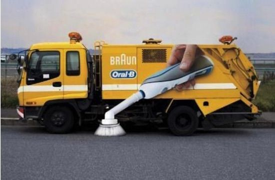 A yellow-colored garbage truck featuring a dynamic 3D mobile billboard on its side, promoting the Oral-B brand. The eye-catching ad portrays a person holding Oral-B's innovative toothbrush, which appears to pop out of the billboard and clean the road, resembling a mop. The unique visual captures attention and highlights the effectiveness of Oral-B's new toothbrush