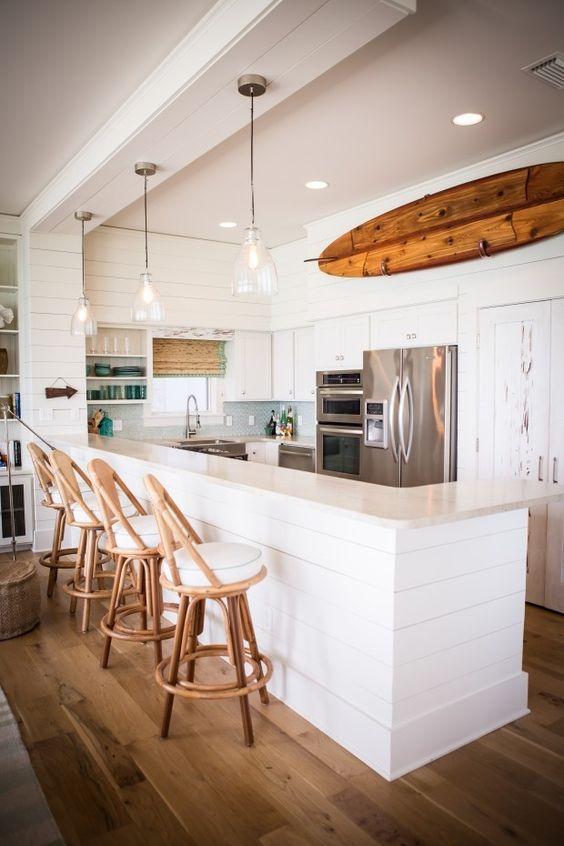 Cozinha com toque rústico, bancada e armários de madeira branca, eletrodomésticos de inox, prateleira grande decorativa amadeirada, piso de madeira, luminárias pendentes transparente e bancos com estrutura de madeira e estofado branco.