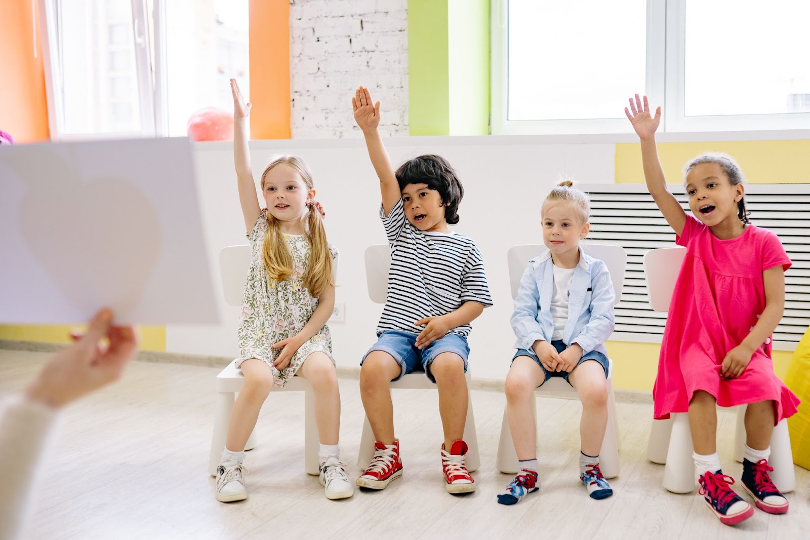 Children sitting on stools in classroom raising their hands in the air.