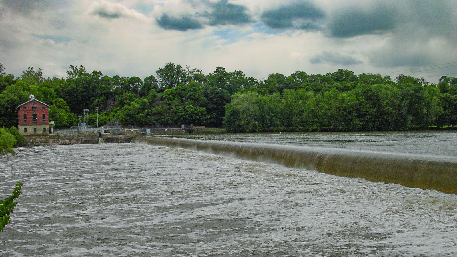 A river flows over a weir with a 2 story building on the far shore. 