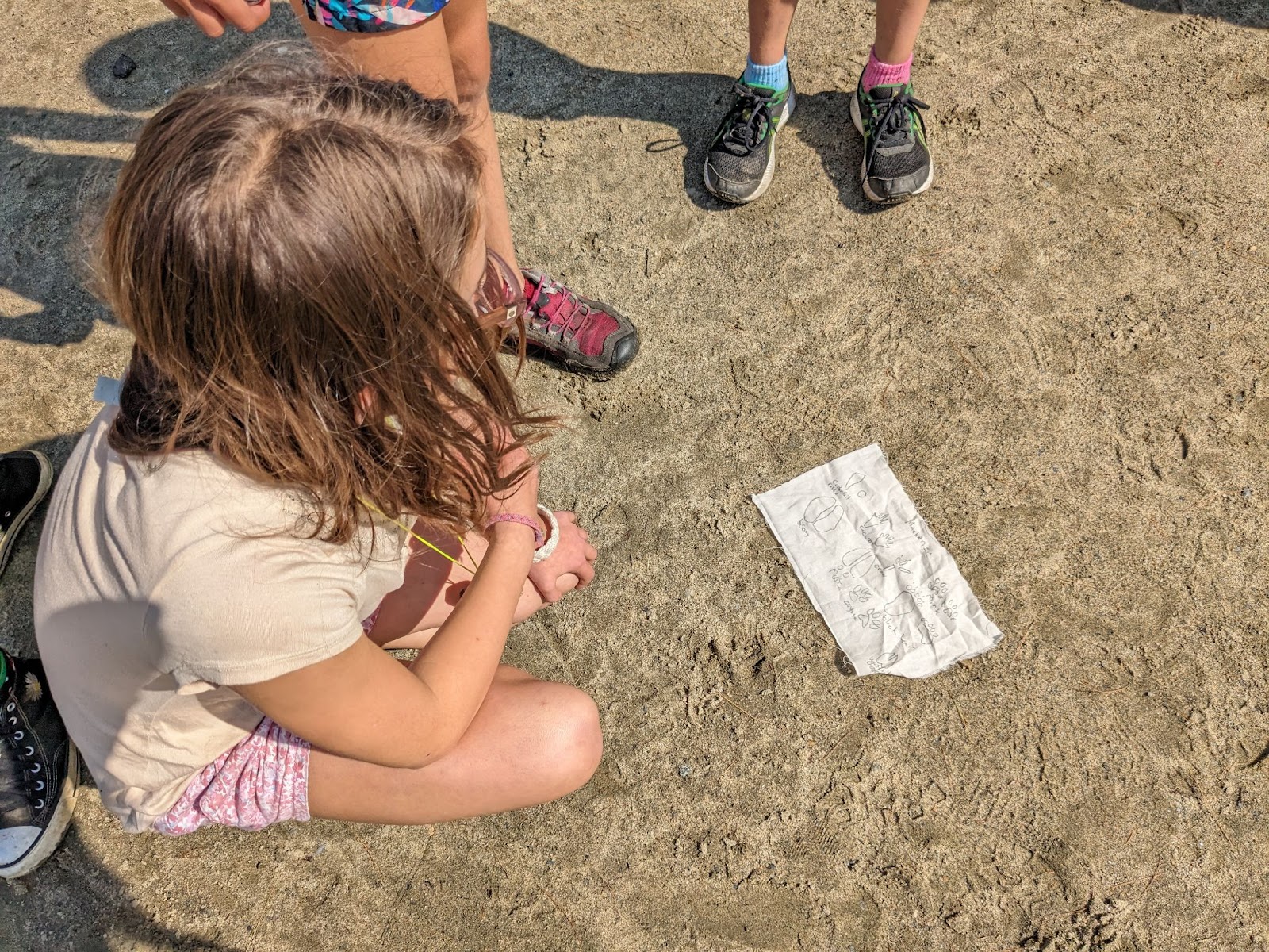 One kid kneeling infant of piece of paper laying on sand at Storrs Pond surrounded by other kids