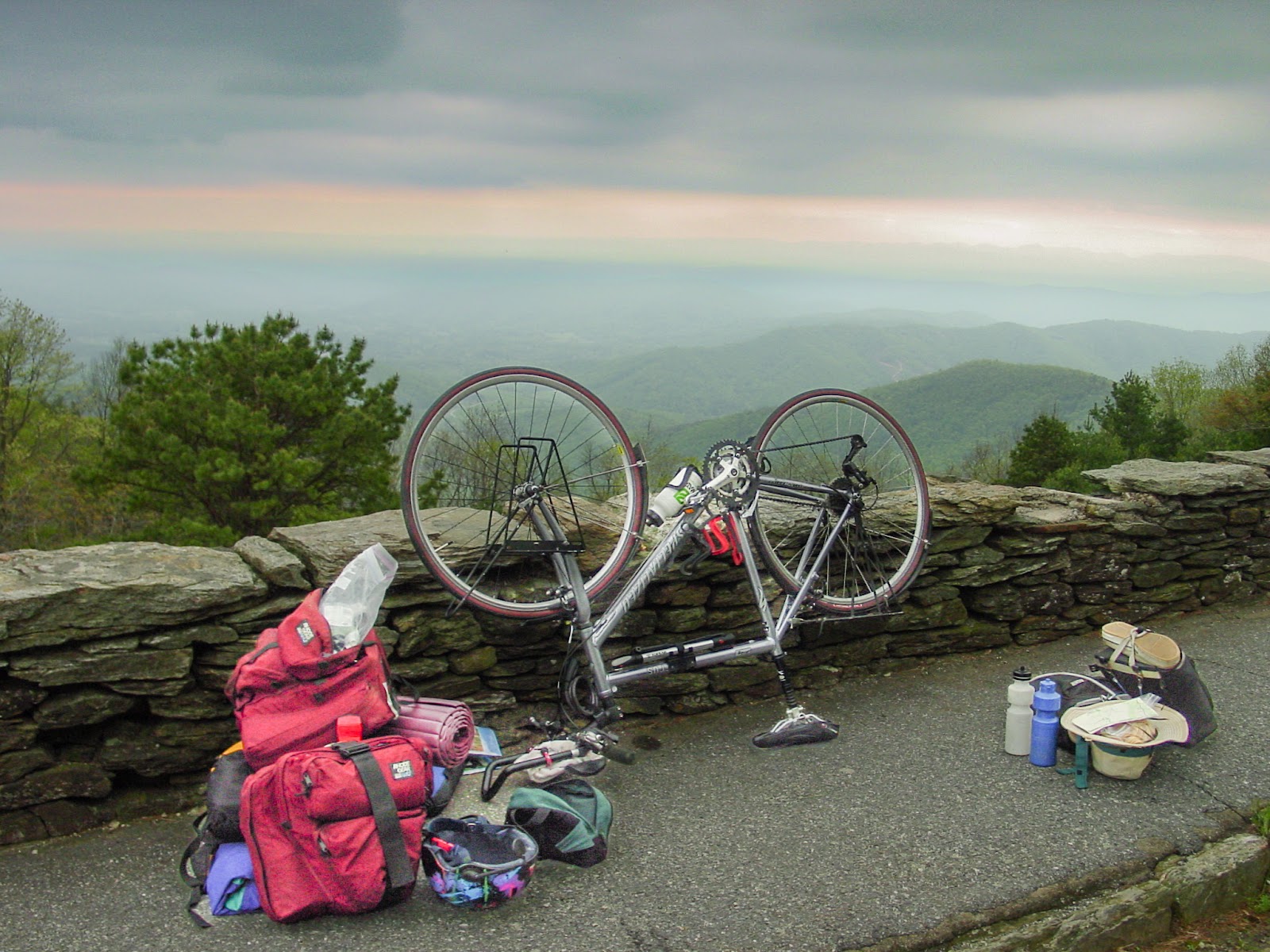 A bike turned upside down with gear spread around it leaning on a rock wall overlook. The sky has a yellow streak and is cloudy and dark. 