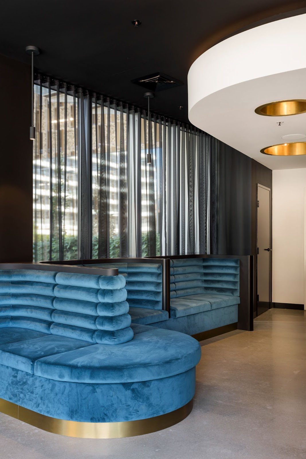 Modern interior with a sleek teal velvet booth seating, black sheer curtains, and gold-accented lighting fixtures.