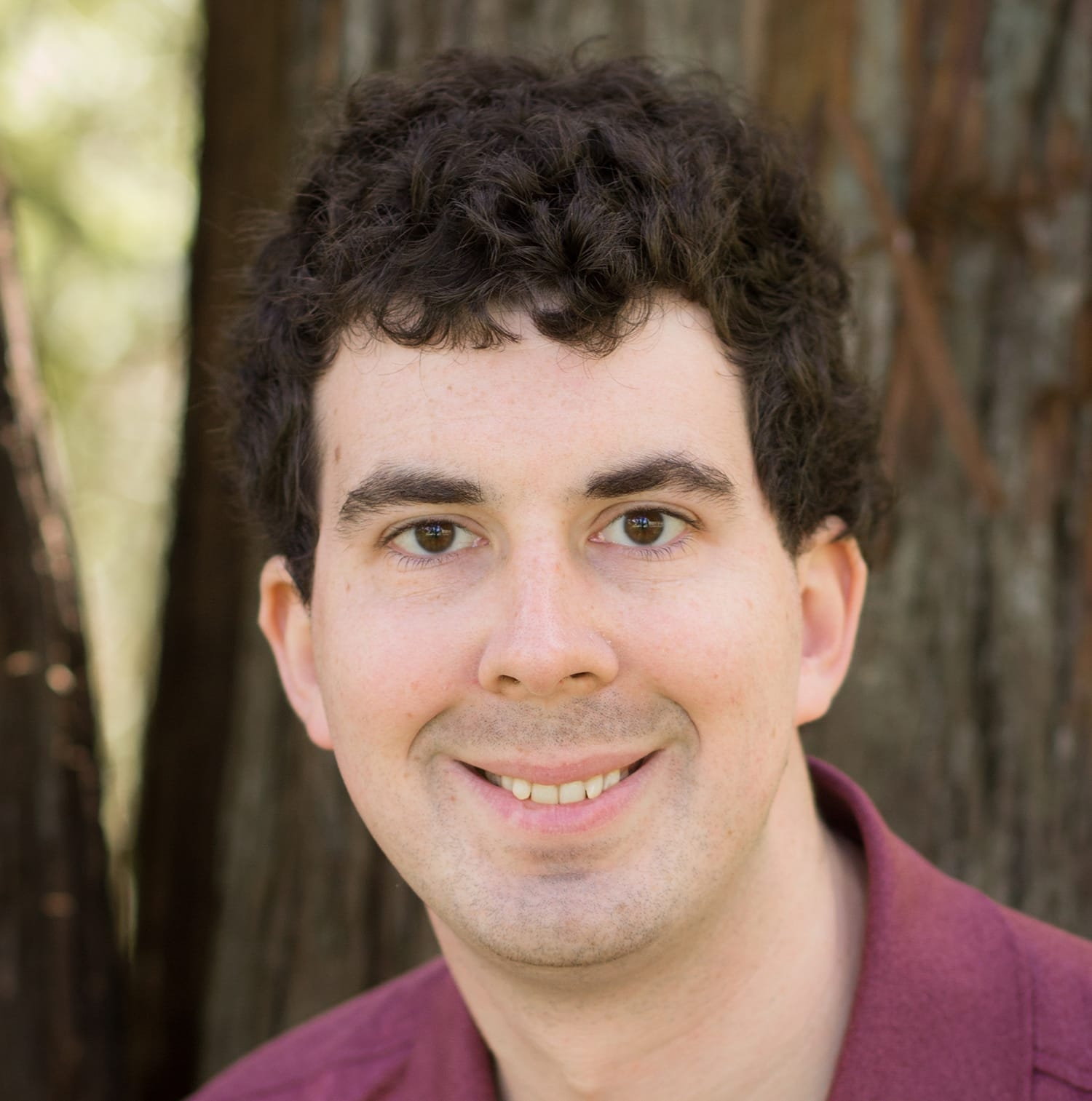 Smiling man wearing burgandy shirt, tree in background