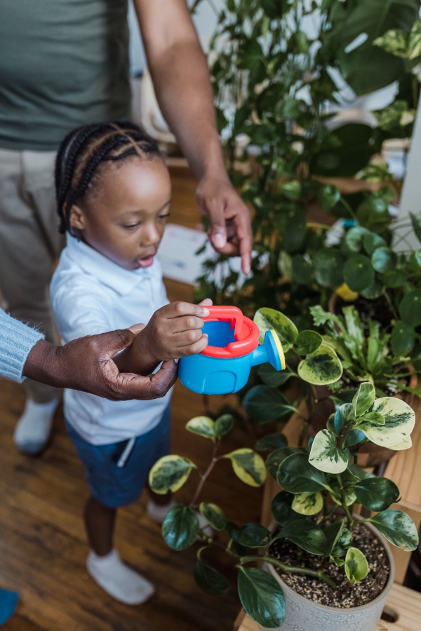 A young child watering a plant with a blue, toy watering can.