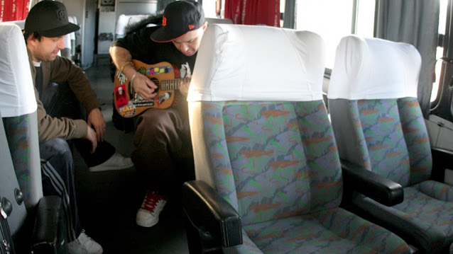 Marcão sentado num ônibus, possivelmente da banda, ao lado de Chorão com violão em mãos enquanto parece ensaiar uma canção.