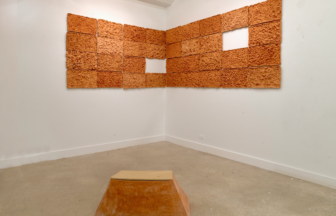 Image: installation shot of In the Moment. A corner of the exhibition featuring a large orange rectangle composed of smaller rectangles with varying textures and colors. There are two rectangular holes in the grid. Image courtesy of the artist.