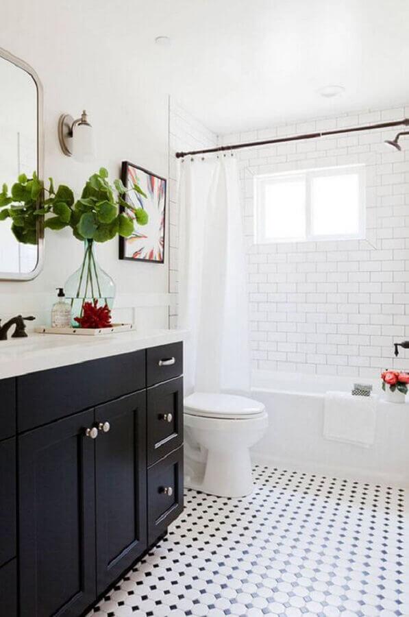 Banheiro com armário preto com bancada branca, área do box com banheira e azulejo subway tiles branco, demais paredes pintadas de branco e piso branco com bolinhas pretas.
