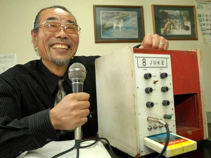 Daisuke Inoue, karaoken keksijä, pitelee mikrofonia alkuperäisen 8 Juken karaokekoneensa kanssa.