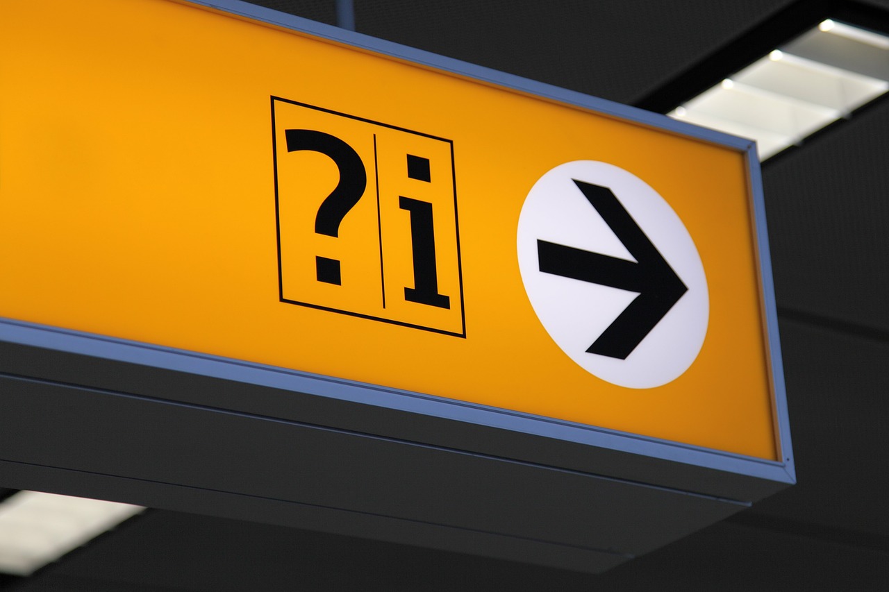 एक संकेत जो किसी हवाई अड्डे पर छत से लटका हुआ है, जिसमें प्रश्न चिह्न, अक्षर i, और एक तीर की ओर इशारा करता है।