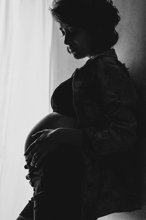 Side view of black and white crop calm pregnant woman in crop top and jacket standing near wall at home and touching tummy