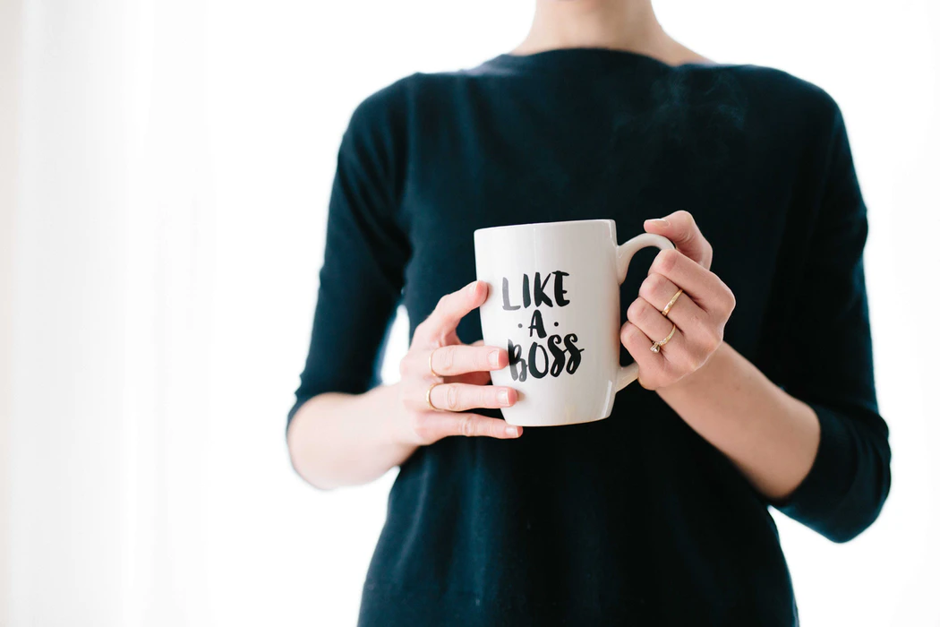 Close up of a woman standing in a black dress holding a white mug with text on it that says ‘like a boss’.