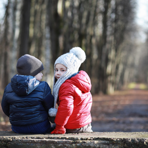 Back view of two children wearing puffer coasts and beanies sitting.  One is looking back at the photographer taking the photo.