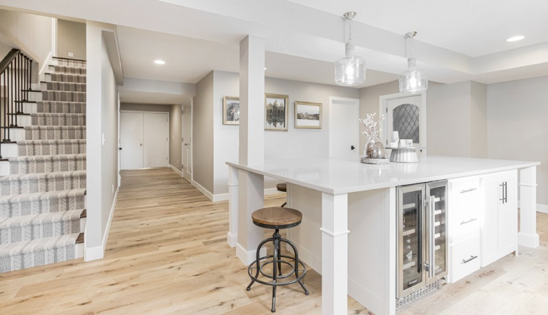 A remodeled basement with wood floors, carpeted stairs, and a full kitchen. The kitchen has a large island with a built-in wine cooler and pendant lights hanging above it.