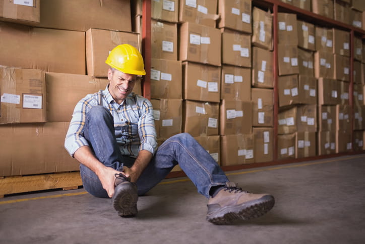 a man in a yellow construction hat is leaning against a wall of boxes as he clutches his ankle in pain after a workplace injury.