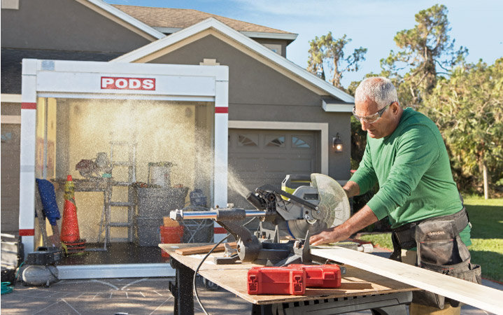 A man is using a miter saw to cut a long piece of wood for his camper renovation. There is a PODS storage container in the background that is being used to store tools and other remodeling supplies.