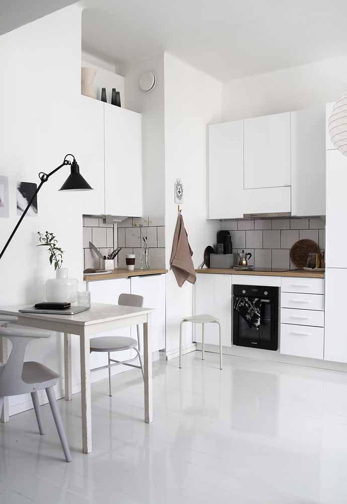 Cozinha com armários brancos, bancadas em tom amadeirado, mesa com cadeira brancas, piso porcelanato branco e utensílios e luminária pretos.