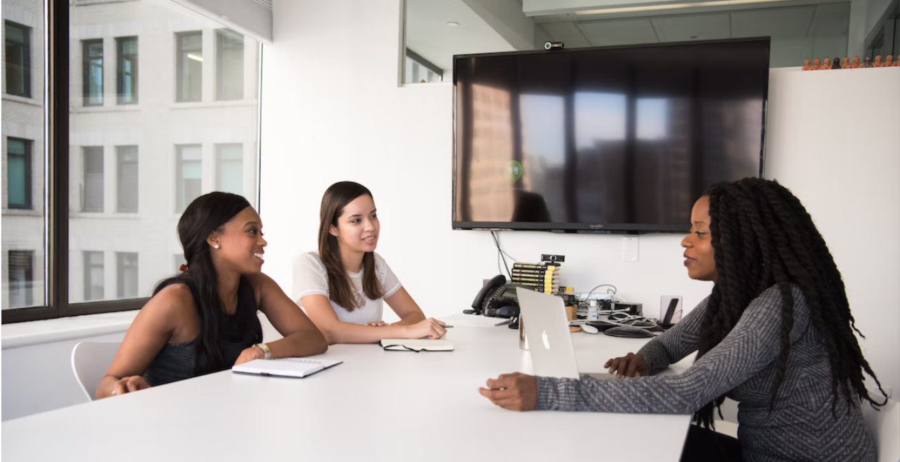 three diverse women sitting around a board table, two on one side are interviewing the other one across the table