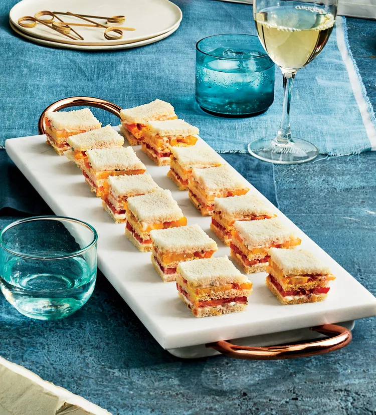 A white marble serving tray with neatly arranged bite-sized sandwiches, layers of bread and fillings, accompanied by glasses of white wine and sparkling water on a blue tablecloth.