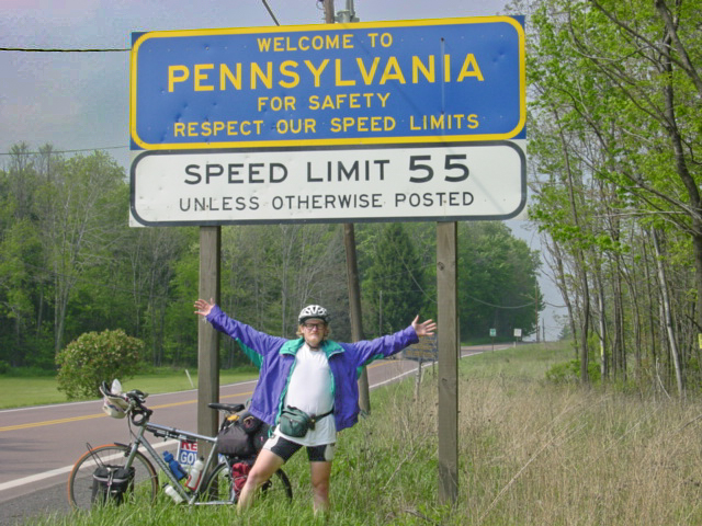 Man with bike, his arms apart under the sign that says: WELCOME TO PENNSYLVANIA FOR SAFETY REPECT OUR SPEED LIMITS. SPEED LIMIT 55 UNLESS OTHERWISE POSTED.