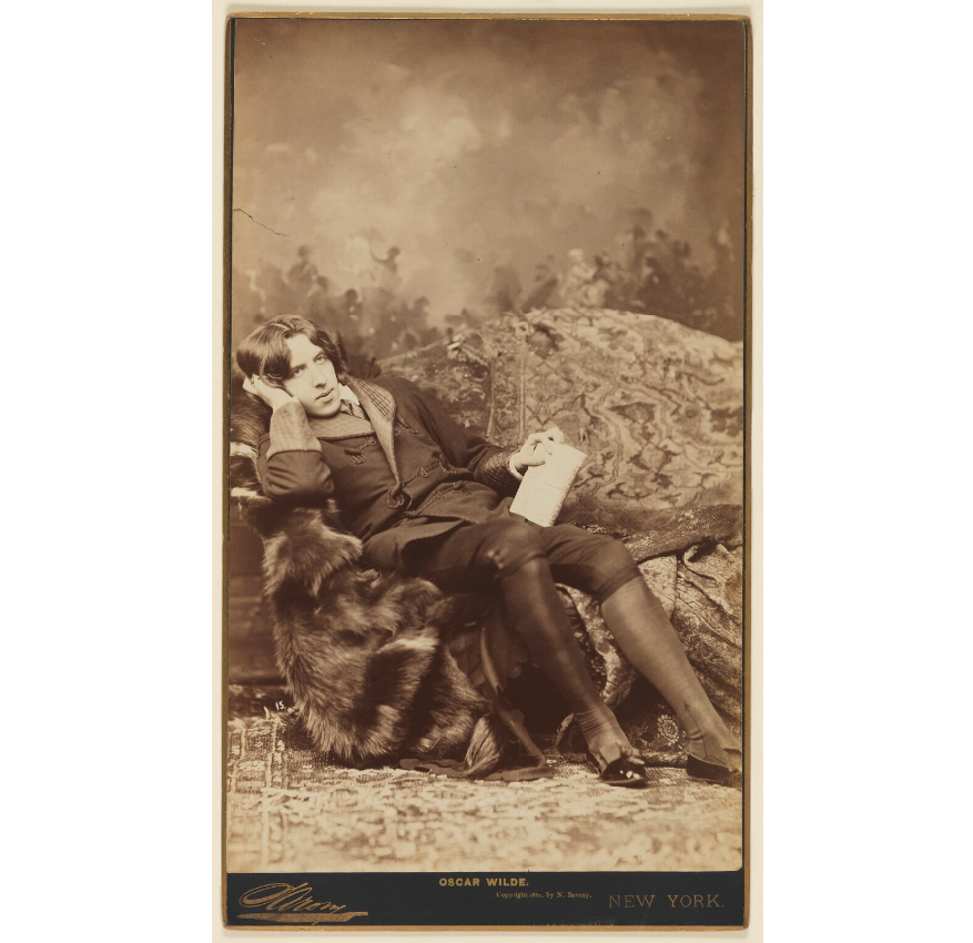 Sepia toned photograph of Oscar Wilde,  by Napoleon Sarony, 1882. Wilde is shown wearing aesthetic dress and lounging on a seat covered with an animal pelt. 