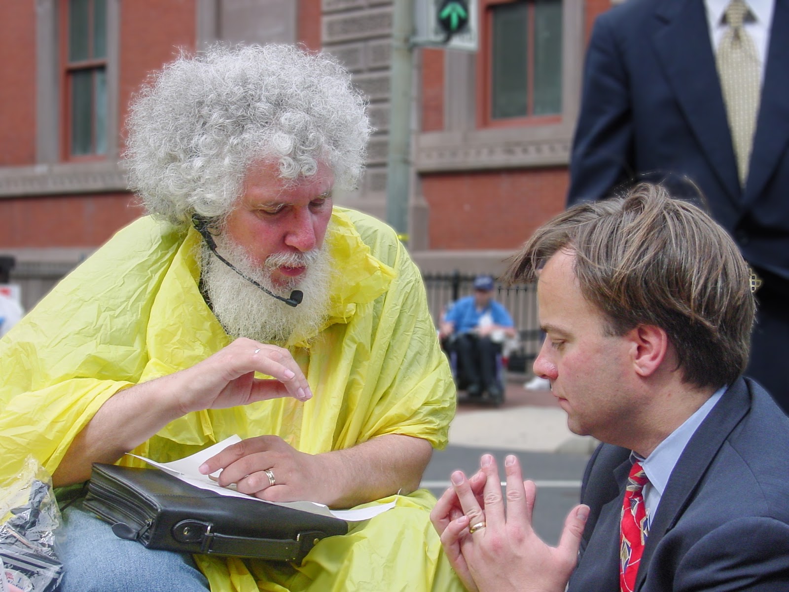 Man with white hair and beard in a yellow rain poncho speaks with a young man in a blue suit.