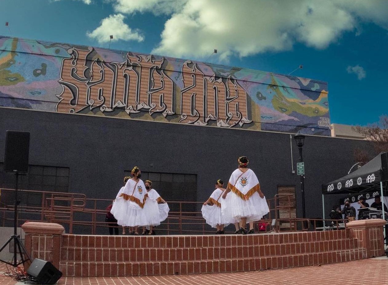 A picture of a group of dancers on stage in white dress and the backdrop said Santa Ana