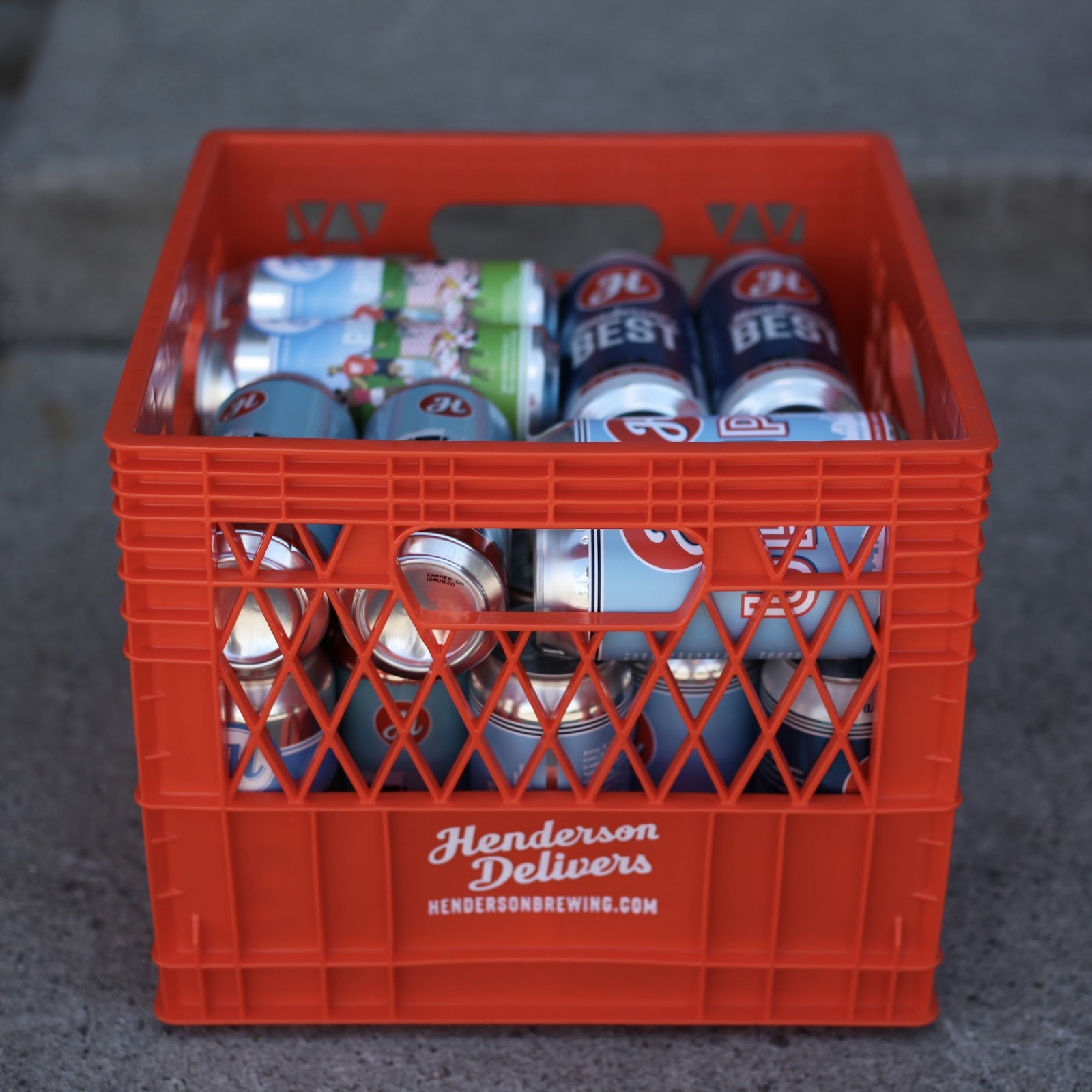 A bright orange milk crate that says 'Henderson Delivers' on it, filled with cans of delicious Henderson beer