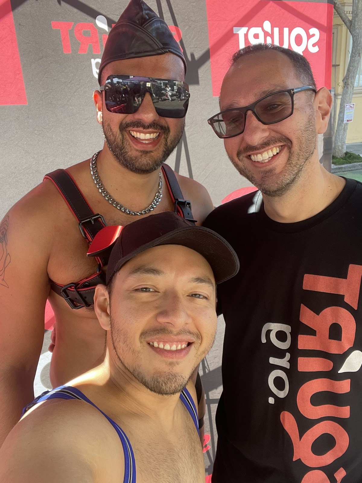 Three gay males posing for a selfie with two wearing gay leather harnesses and one wearing a black Squirt.org logo t-shirt