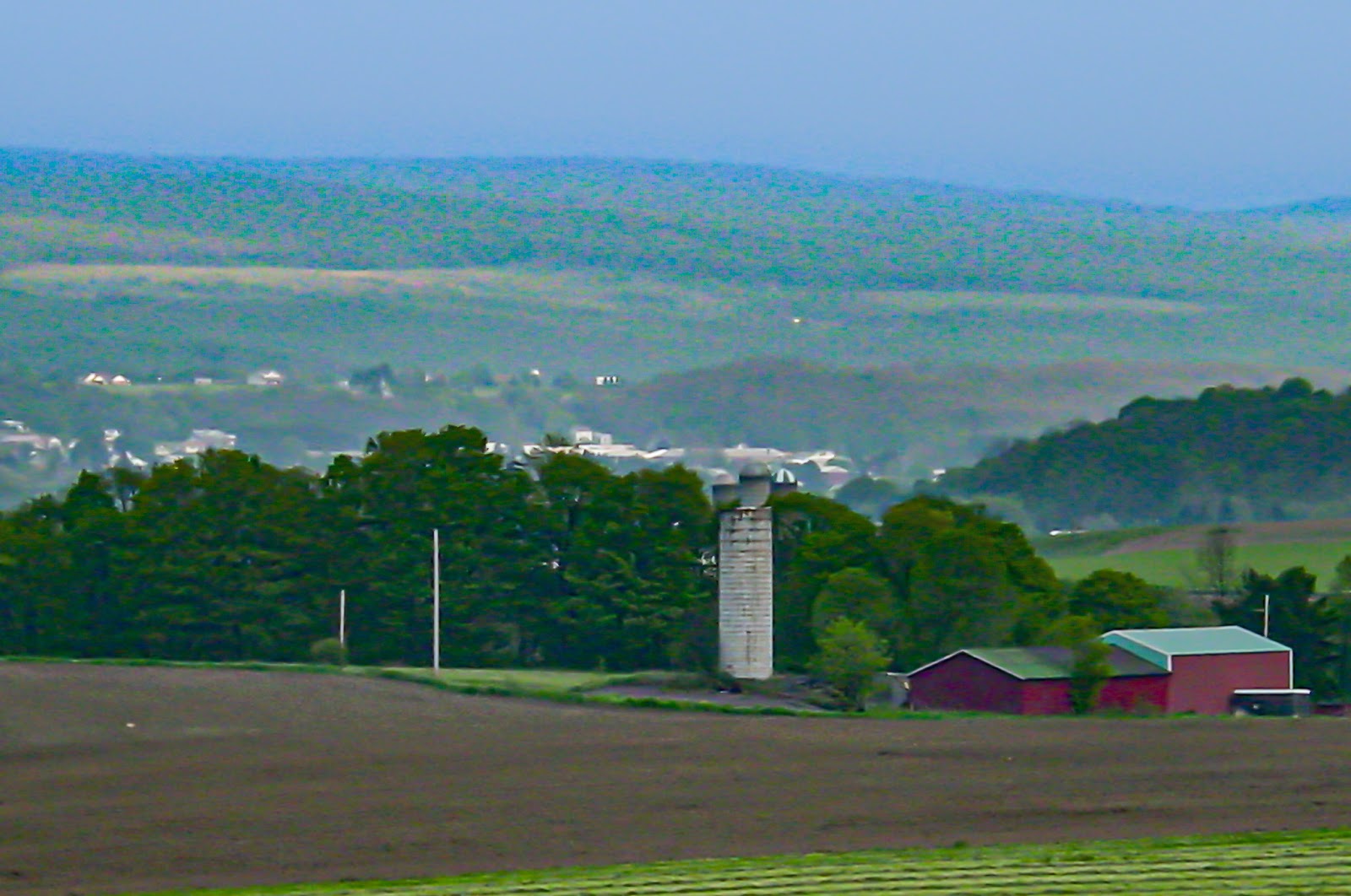 A plowed field, silo and red barn  in the foreground with a community in the distance and low hills. 