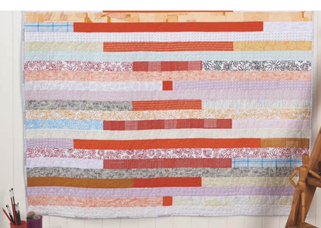 This is an image of a quilt hanging on a wall.  It is made up of horizontal striped pieces.  Every other stripe is a single fabric, and the alternate strip is pieced from several different fabrics.  The fabrics are generally pastel prints, with a few bright orange & red fabrics used in the piecing.  